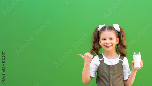A little girl drinks cow's milk and points her finger at an advertisement on an isolated green background. Calcium for the child's body, copy space.