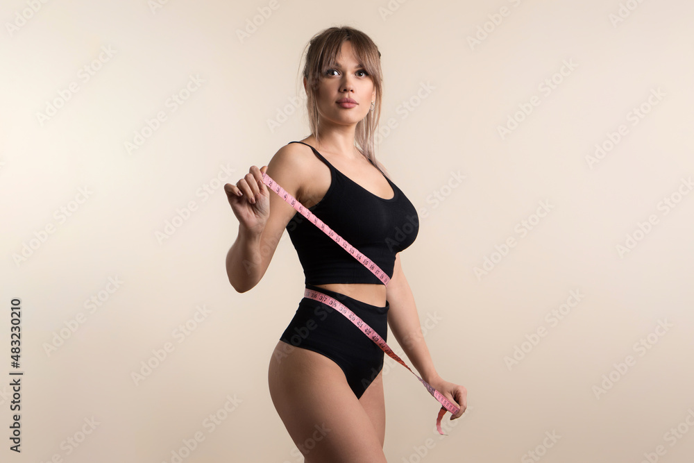 Obraz premium Slim healthy girl in black underwear with measuring tape at waist. Fit Woman poses in high waist panties and top. Perfect female body. Fitness, Diet or body care concept. Female shows her flat belly