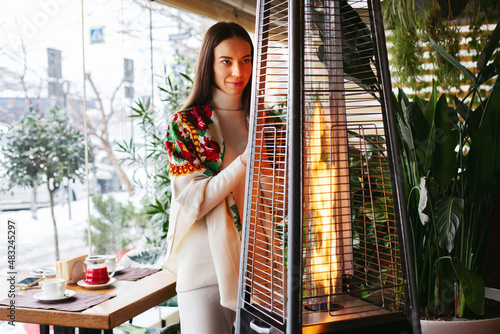 A young brunette girl in a cafe is warming herself near the fireplace, resting