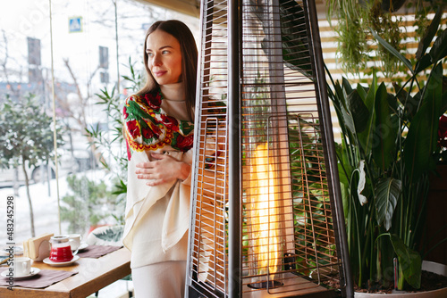 A young brunette girl in a cafe is warming herself near the fireplace, resting