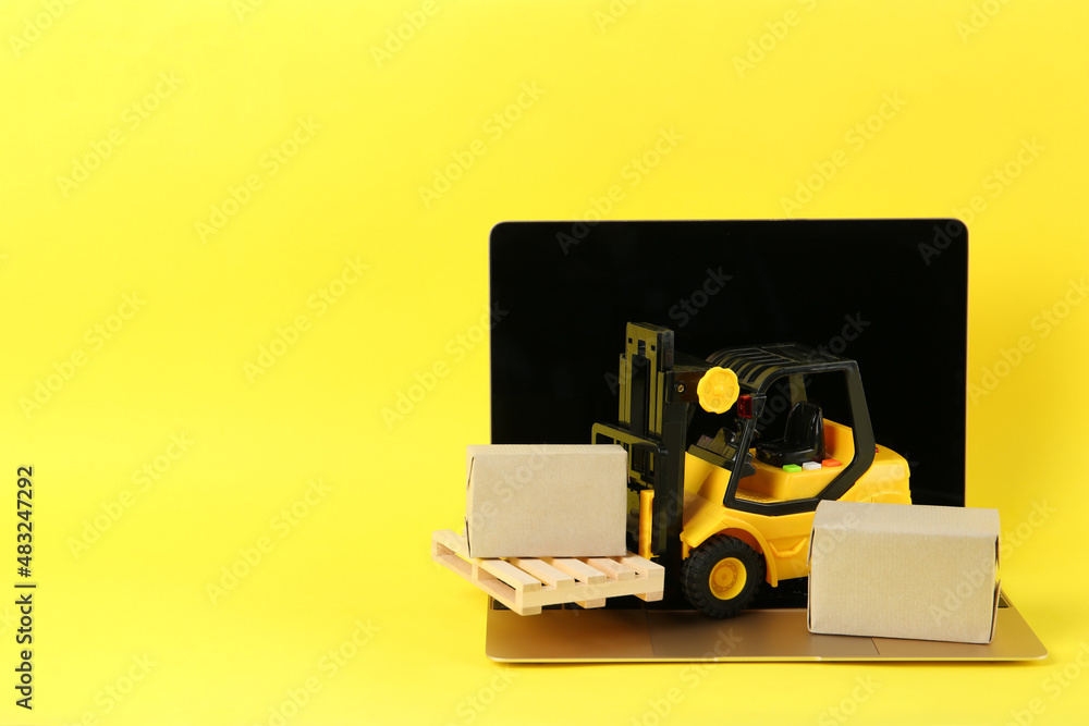 Laptop, toy forklift with wooden pallet and boxes on yellow background, space for text