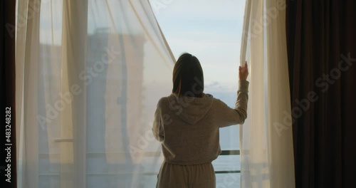 Back view of young woman closing curtains in apartment. Unrecognizable girl traveler enjoying evening weekend relaxing at home alone close-up. End of the day