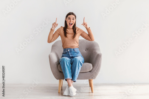 Overjoyed millennial woman sitting in armchair, pointing fingers up, shouting in excitement, offering space for your ad