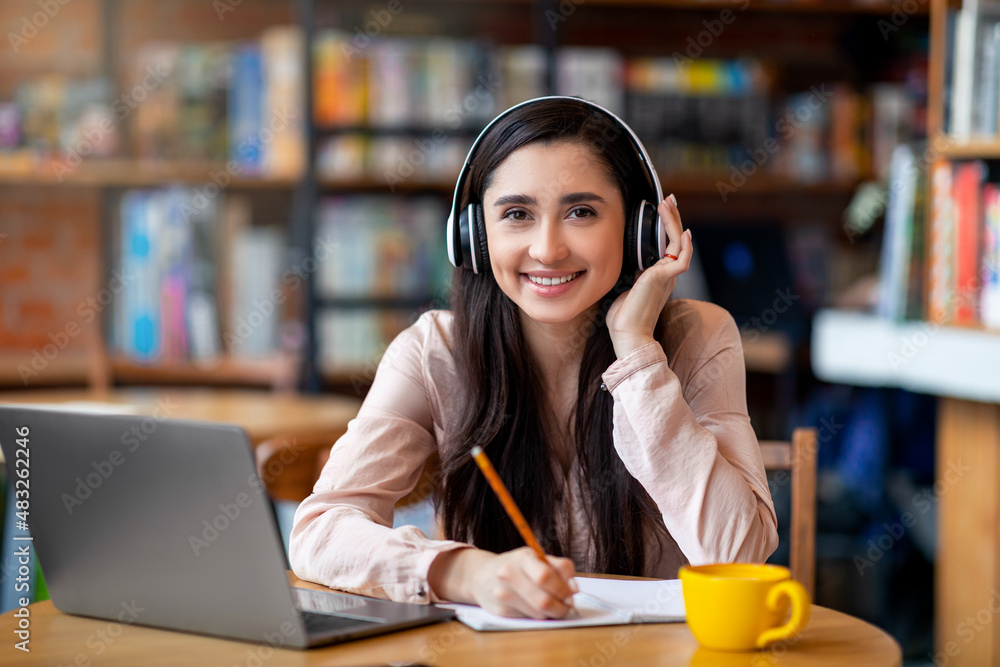 © Prostock-studio - Cheerful latin student in wireless headphones studying online while sitting in cafe, using laptop and taking notes