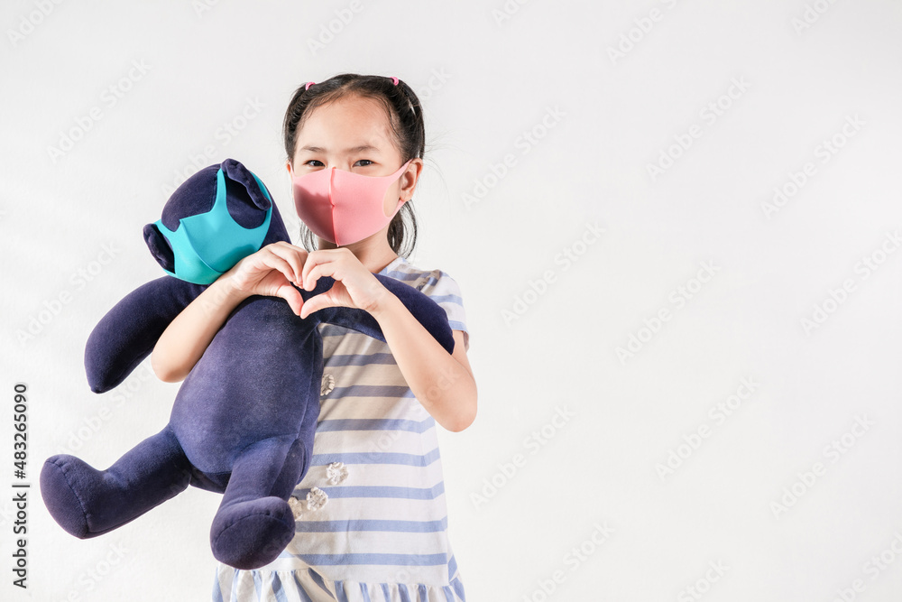 Positive Asian little toddler girl wearing pink mask holding a toy bear shows heart shape gesture love and support. Coronavirus outbreak stays at home concept. White background copy and space idea Positive Asian little toddler girl wearing pink mask holding a toy bear shows heart shape gesture love and support. Coronavirus outbreak stays at home concept. White background copy and space idea