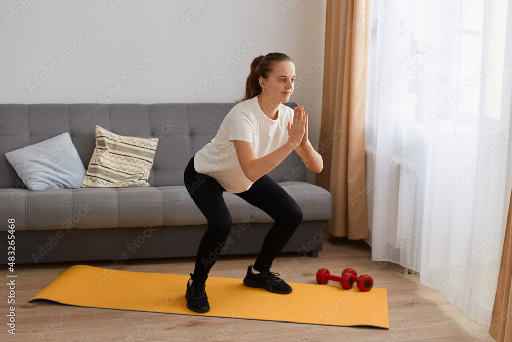 Portrait of athletic woman with ponytail wearing black leggins and white t shirt doing sport exercises on yoga mat, doing sits-up and keeping palms together, or practicing yoga position.