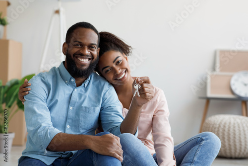 Happy young black couple hugging and showing keys to new apartment with cardboard boxes, looking at camera