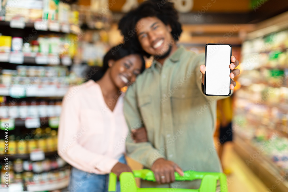 African American Buyers Couple Showing Smartphone Screen Standing In Supermarket