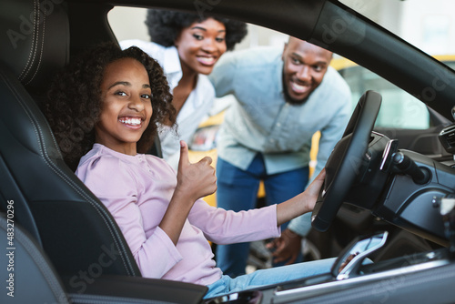 Cute black girl sitting inside new car, showing thumb up gesture, recommending auto dealership with her parents