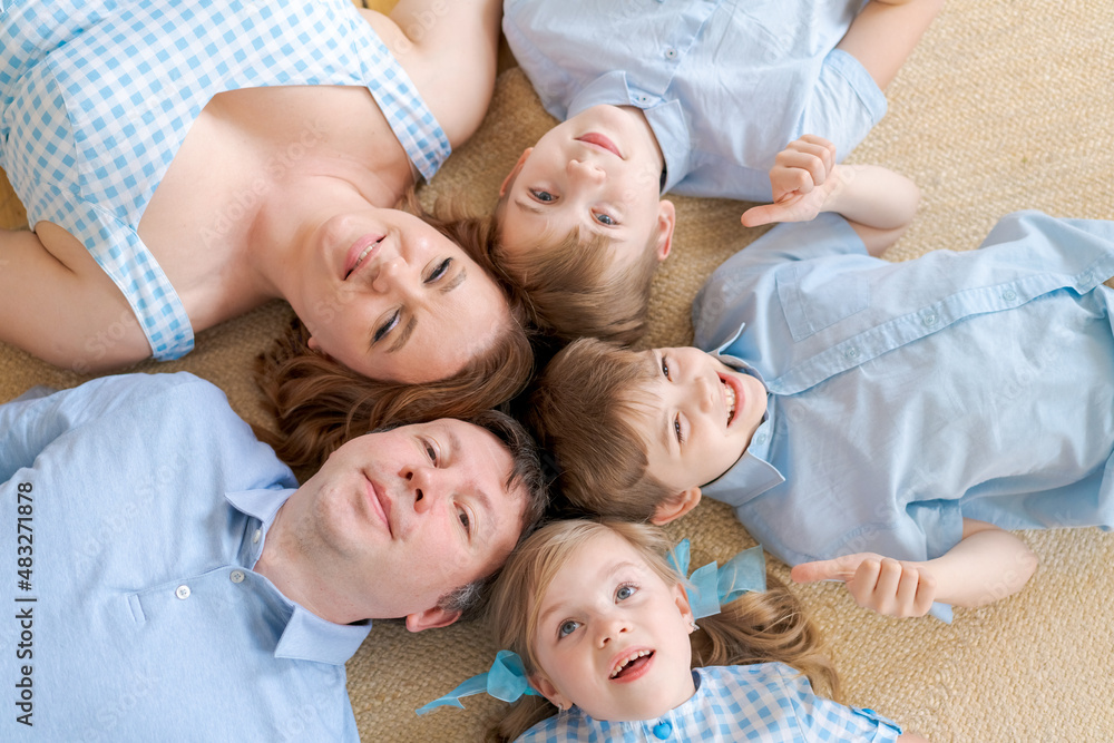 Portrait happy caucasian family posing on floor house, lying on carpet and approaching, smiling cheerfully, top view. Positive european father, mother and daughter and two brothers hugging