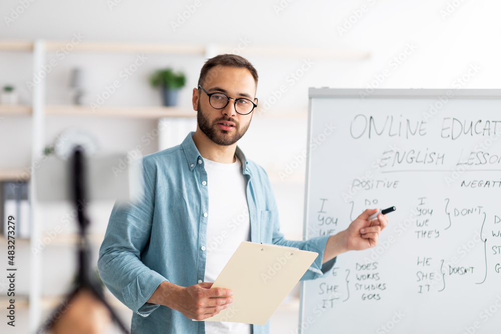 Young male teacher pointing at blackboard with English grammar rules, conducting online lesson from home Young male teacher pointing at blackboard with English grammar rules, conducting online lesson from home