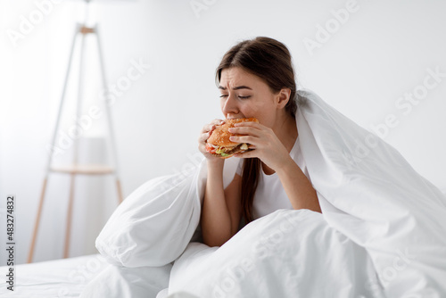 Hungry after diet, starving stressed caucasian millennial woman eating burger in bed photo