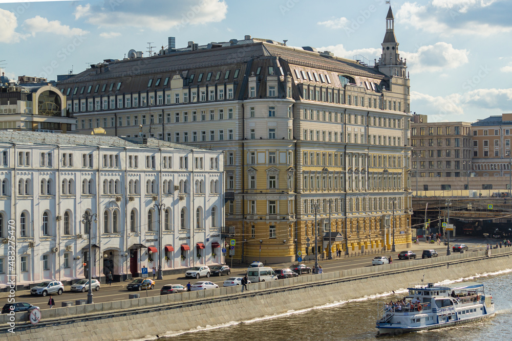 Fototapeta premium Moscow, Russia - June 16, 2019: View of Hotel Baltschug Kempinski on Raushskaya Embankment. Pleasure boat sails along Moscow River in direction of Stalin's skyscraper on Kotelnicheskaya Embankment.