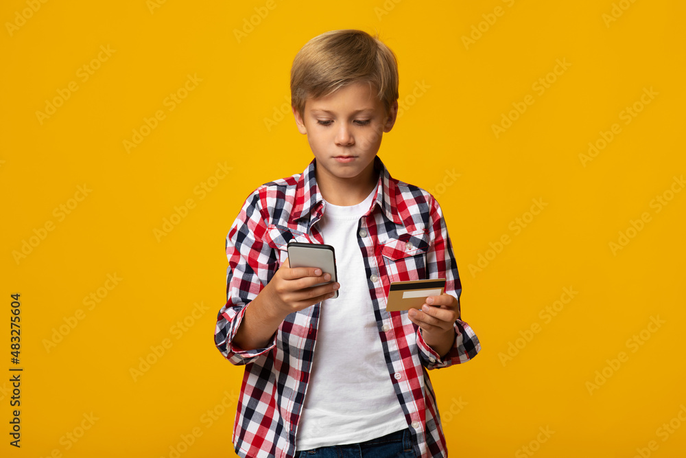 Calm busy caucasian teen boy pupil with credit card typing on phone, isolated on yellow background Calm busy caucasian teen boy pupil with credit card typing on phone, isolated on yellow background