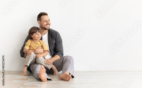 Happy caucasian father carrying little son, sitting together on floor and looking aside at empty space, white background