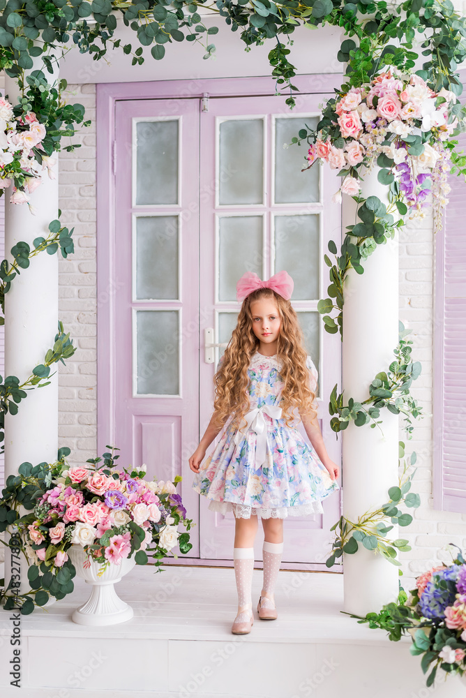 girl who looks like a doll is standing on the veranda of a house decorated with flowers. A child with long blonde curls and a big pink bow on her head