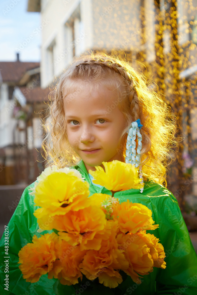 Cute little blonde girl in green raincoat with yellow flowers on a green lawn under rain drops in a summer sunny day near rural house in a village Cute little blonde girl in green raincoat with yellow flowers on a green lawn under rain drops in a summer sunny day near rural house in a village