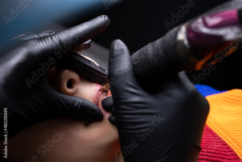 a tattoo artist in black sterile gloves holds a tattoo machine and puts a tattoo on the lower lip of the model