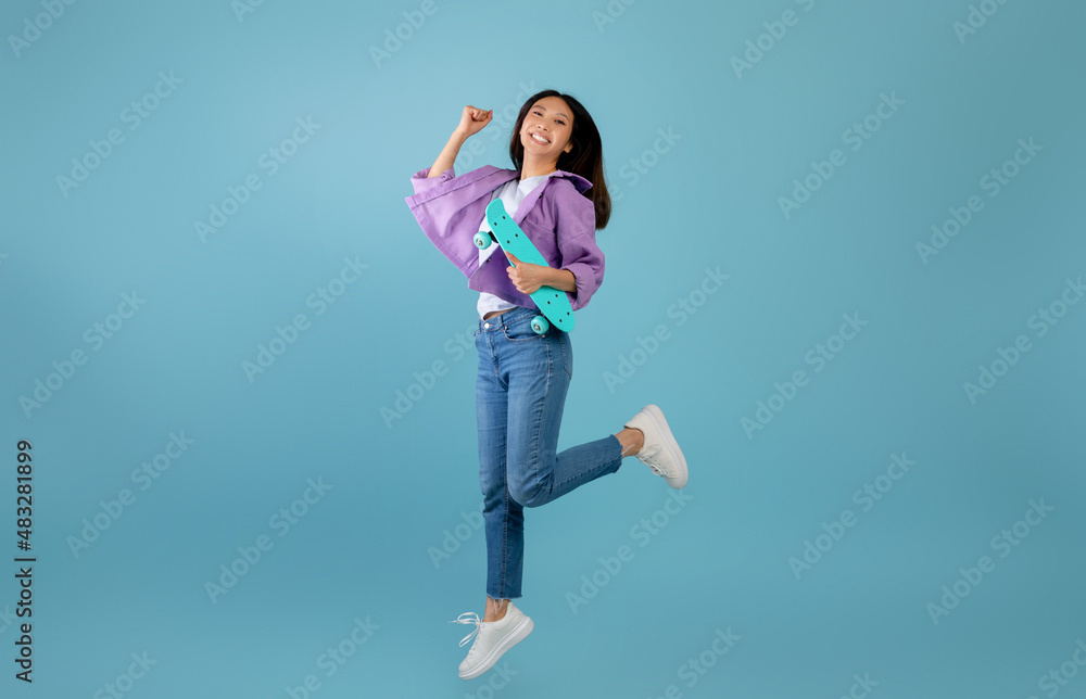 Teenage culture. Playful asian lady with skateboard jumping and having fun over blue studio background