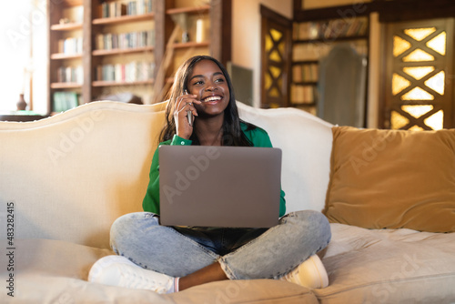 Smiling young black woman having conversation on cellphone, working on laptop in at home
