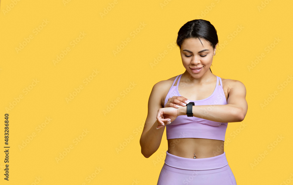 Happy african american lady using smartwatch to monitor progress, checking her performance, yellow background Happy african american lady using smartwatch to monitor progress, checking her performance, yellow background