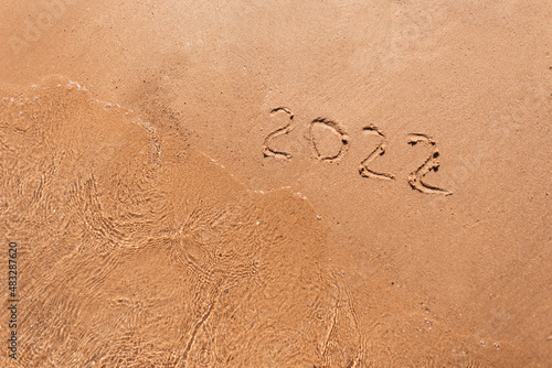 Happy New Year 2022, lettering on the beach with wave and clear sea. Numbers 2022 year on the sea shore, message handwritten in the golden sand on beautiful beach background. New Years concept.