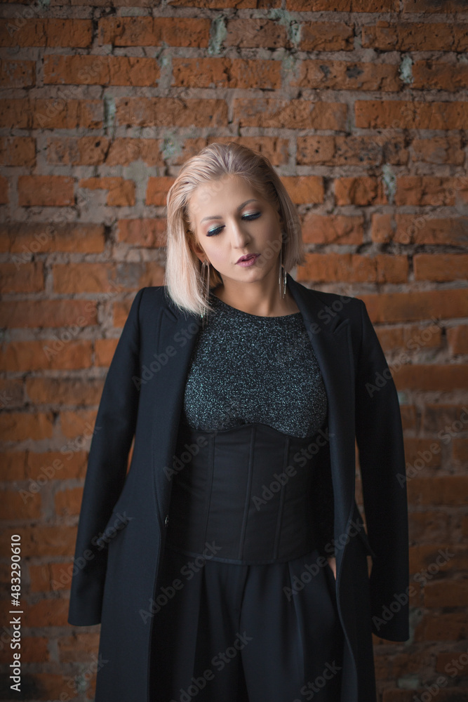 blonde girl in a shiny jacket, a black corset and a coat is standing near a brick wall blonde girl in a shiny jacket, a black corset and a coat is standing near a brick wall