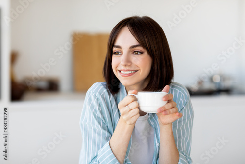 Morning coffee. Portrait of young lady with mug in hands, sitting in kitchen interior and looking aside, free space