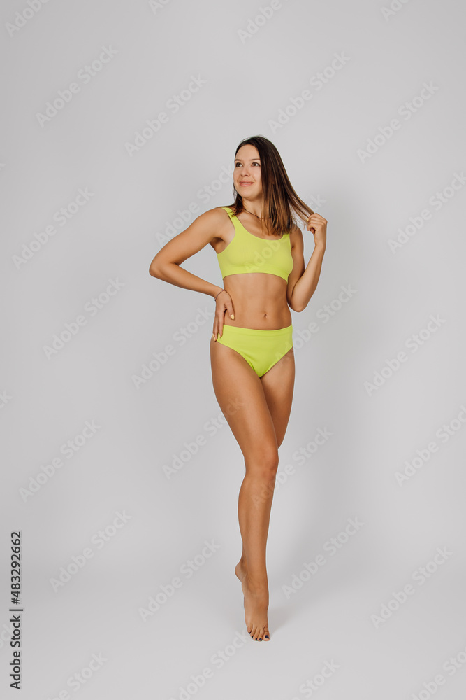 Brunette shows off slender figure. Young beautiful Caucasian woman in yellow swimsuit or bikini poses on light isolated background, stands in elegant pose and holds her hair with her hands. Brunette shows off slender figure. Young beautiful Caucasian woman in yellow swimsuit or bikini poses on light isolated background, stands in elegant pose and holds her hair with her hands.