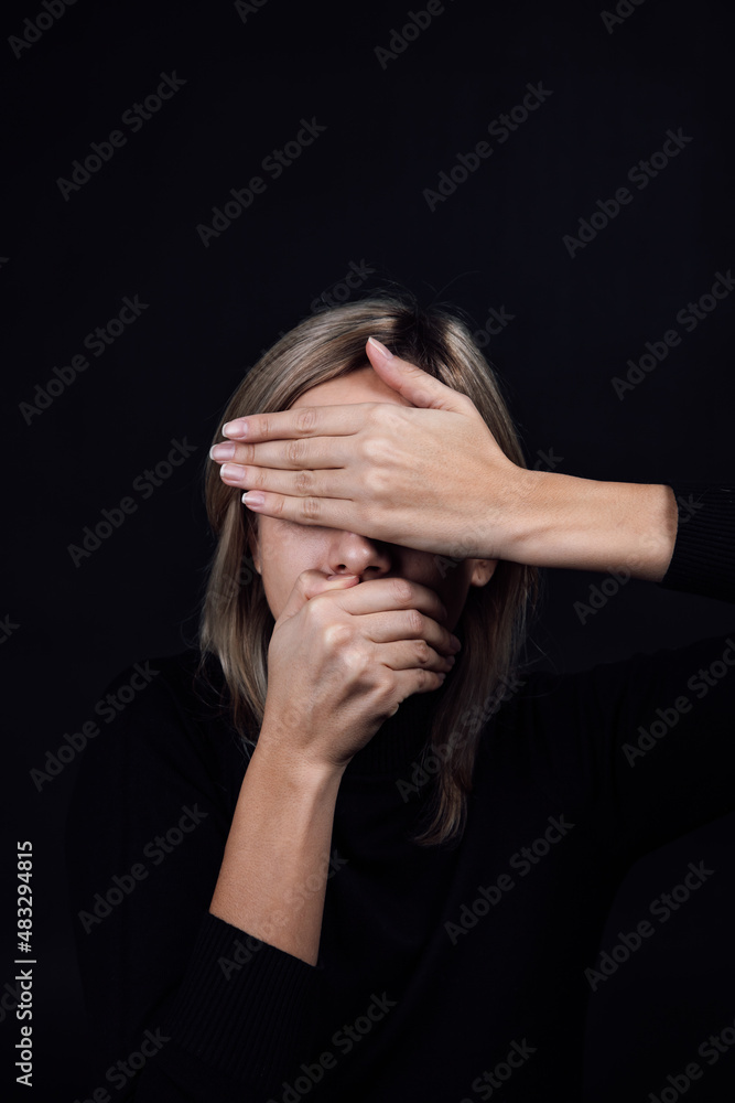 Oppressed woman with hands hiding eyes and mouth wearing black blouse on black background. Victim of physical and psychological abuse. Gaslighting. Relative aggression. Copy space. Oppressed woman with hands hiding eyes and mouth wearing black blouse on black background. Victim of physical and psychological abuse. Gaslighting. Relative aggression. Copy space.