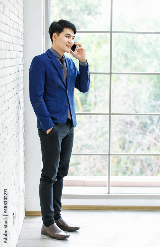 Portrait studio shot of Asian handsome professional successful male businessman entrepreneur in formal blue suit necktie standing holding smartphone smiling on white brick wall background. Portrait studio shot of Asian handsome professional successful male businessman entrepreneur in formal blue suit necktie standing holding smartphone smiling on white brick wall background.