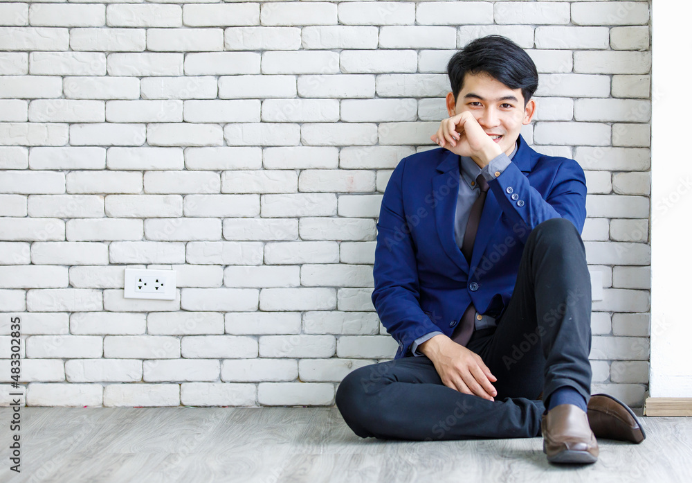 Portrait studio shot of millennial Asian handsome professional successful male businessman entrepreneur in formal blue suit necktie sitting smiling posing leaning on white brick wall background Portrait studio shot of millennial Asian handsome professional successful male businessman entrepreneur in formal blue suit necktie sitting smiling posing leaning on white brick wall background