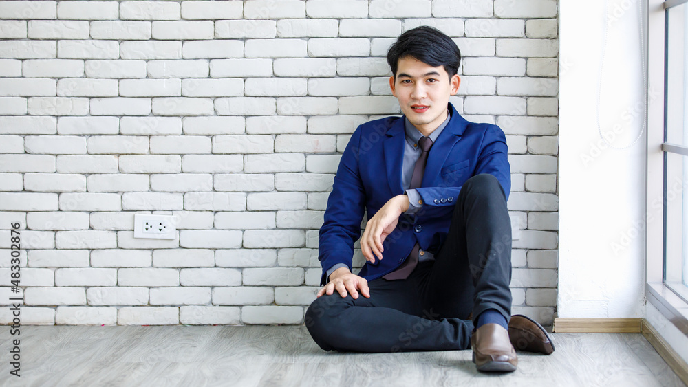 Portrait studio shot of millennial Asian handsome professional successful male businessman entrepreneur in formal blue suit necktie sitting smiling posing leaning on white brick wall background