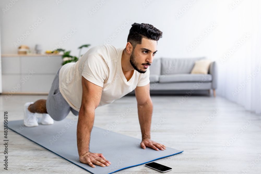 Fototapeta premium Strength workout. Fit young Arab man doing push ups or plank at home, following video tutorial on smartphone, free space