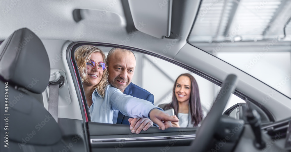 Wife shows her husband features of a new car in the dealership, smiling, while the sales assistant stands aside