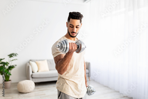 Domestic training concept. Strong young Arab man exercising with dumbbells at home photo