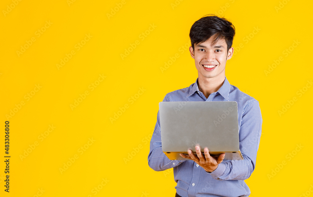 Portrait studio shot of millennial Asian professional successful male businessman employee in formal outfit standing smiling holding laptop computer browsing surfing internet on yellow background Portrait studio shot of millennial Asian professional successful male businessman employee in formal outfit standing smiling holding laptop computer browsing surfing internet on yellow background