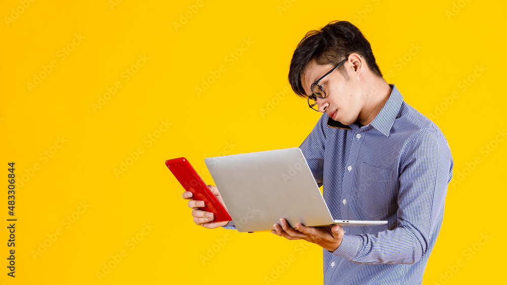 Studio shot of millennial busy stressed overload Asian professional male businessman employee holding laptop computer and tablet in hands on call with customer via smartphone on yellow background Studio shot of millennial busy stressed overload Asian professional male businessman employee holding laptop computer and tablet in hands on call with customer via smartphone on yellow background