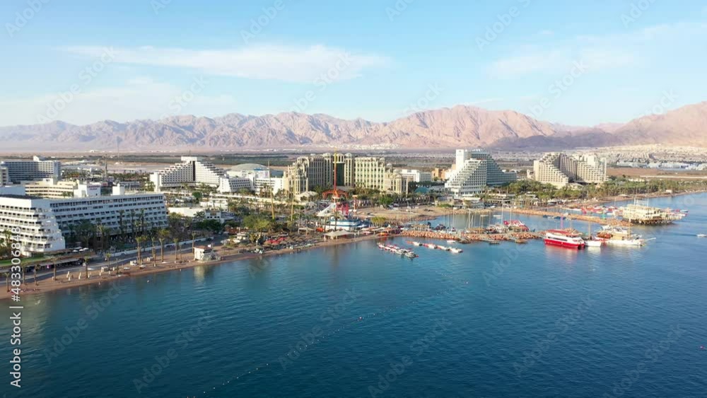 Drone view of Eilat Israel on sunny clear day from the sea featuring boats, luxury hotels and resort area with beach port marina and promenade. The desert and mountains are seen in the background. Drone view of Eilat Israel on sunny clear day from the sea featuring boats, luxury hotels and resort area with beach port marina and promenade. The desert and mountains are seen in the background.