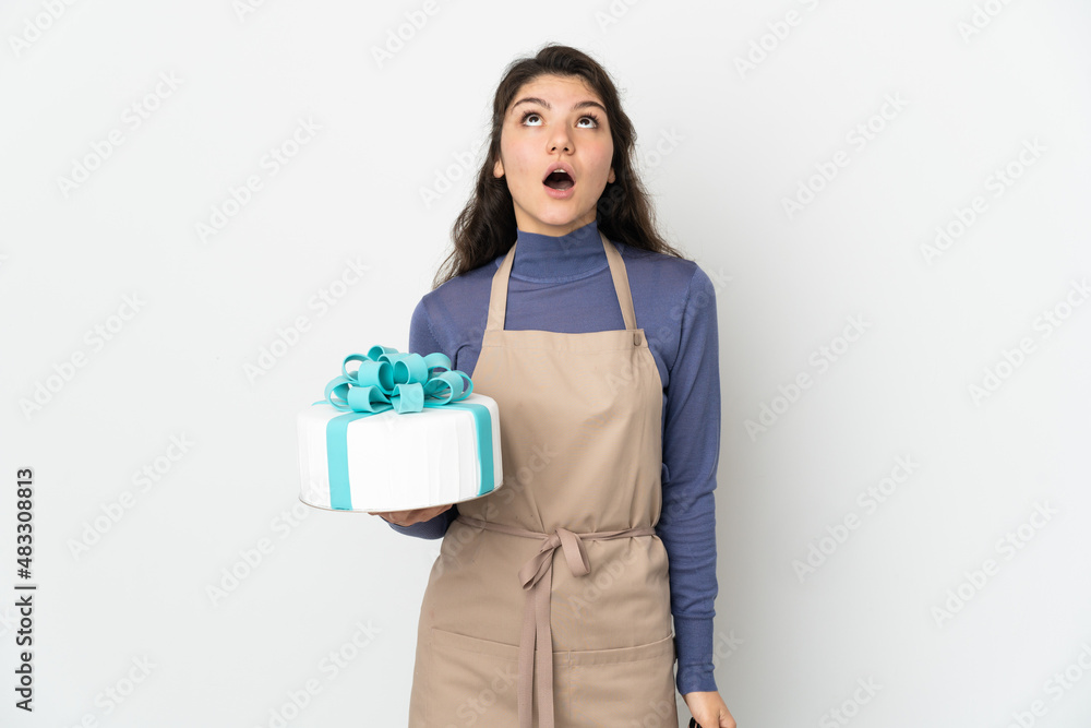 Pastry Russian chef holding a big cake isolated on white background looking up and with surprised expression Pastry Russian chef holding a big cake isolated on white background looking up and with surprised expression