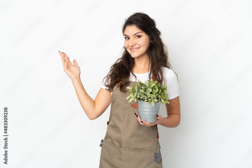 Young Russian gardener girl holding a plant isolated extending hands to the side for inviting to come Young Russian gardener girl holding a plant isolated extending hands to the side for inviting to come