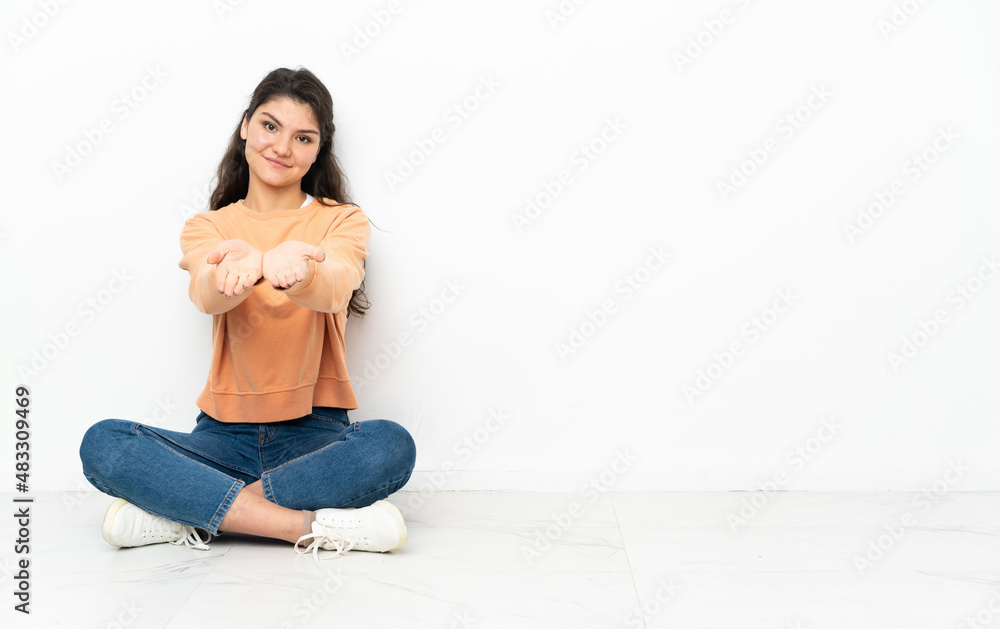 Teenager Russian girl sitting on the floor holding copyspace imaginary on the palm to insert an ad Teenager Russian girl sitting on the floor holding copyspace imaginary on the palm to insert an ad