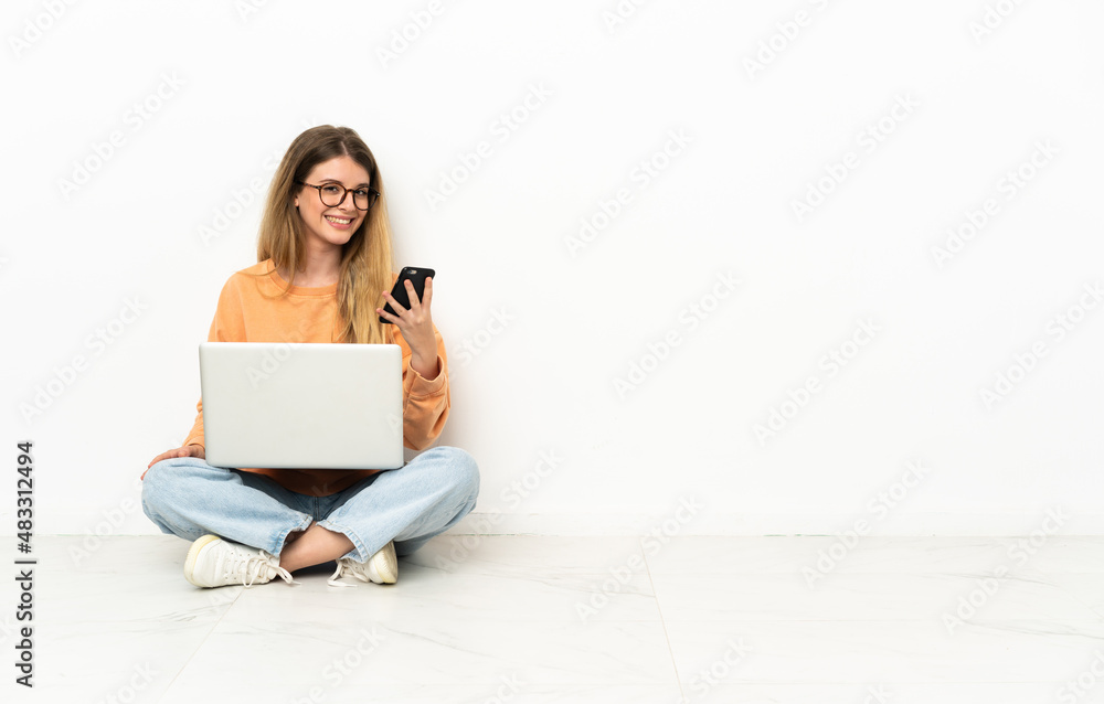 Young woman with a laptop sitting on the floor sending a message with the mobile Young woman with a laptop sitting on the floor sending a message with the mobile