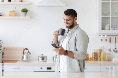 Happy attractive adult caucasian man drinking coffee and reading news at smartphone, chatting in social networks