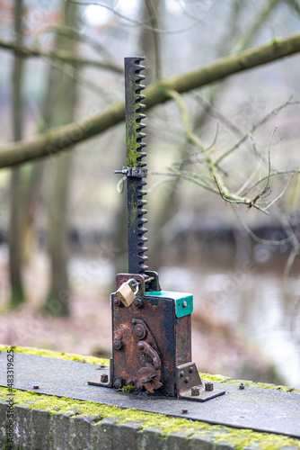 Old and rusty metal part with a cog gear in a floodgate on a bridge, fixed with a padlock so that it is not used, branches of trees and stream in a misty and blurred background in a nature reserve
