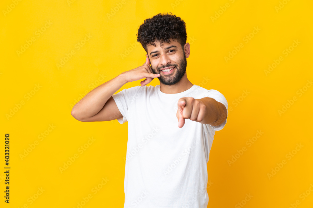 Young Moroccan man isolated on yellow background making phone gesture and pointing front Young Moroccan man isolated on yellow background making phone gesture and pointing front