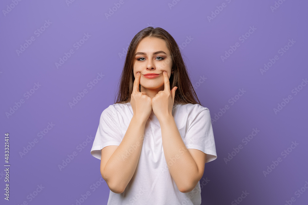 I am not happy with this idea. Studio portrait of gloomy annoyed girl stretching mouth in fake smile with index fingers, standing in blank white t shirt over purple background I am not happy with this idea. Studio portrait of gloomy annoyed girl stretching mouth in fake smile with index fingers, standing in blank white t shirt over purple background