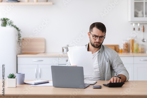 Busy adult caucasian man with beard in glasses counts finances, pays bill and taxes for calculator