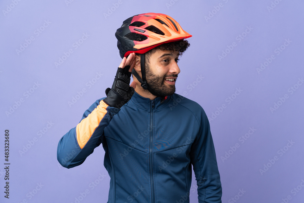Fototapeta premium Young Moroccan cyclist man isolated on purple background listening to something by putting hand on the ear