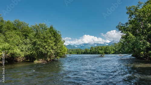 The blue River flows between the banks with lush green vegetation. A picturesque mountain range against the background of azure sky and clouds. Kamchatka